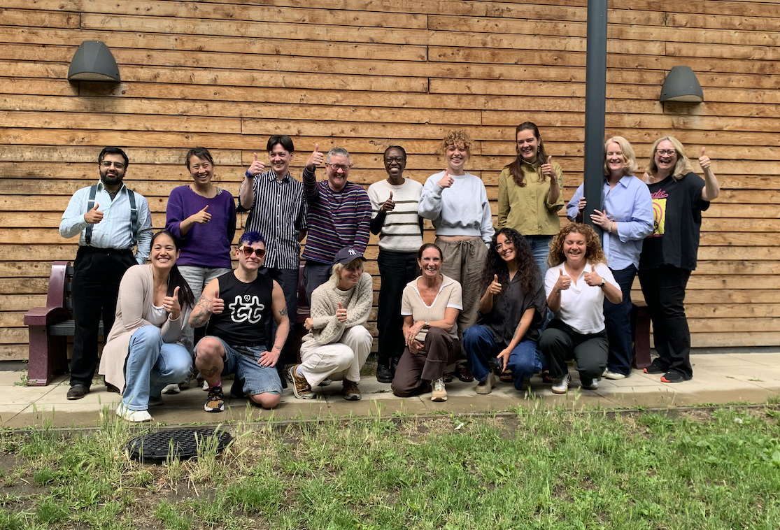 A group of recent participants in the course stand in front of the building where the course is held, some have their thumbs up, all are smiling